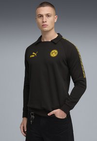 Sort sweatshirt med krave, der har et gult logo og tekst på brystet og langs ærmerne. Lange ærmer og ribbede manchetter.