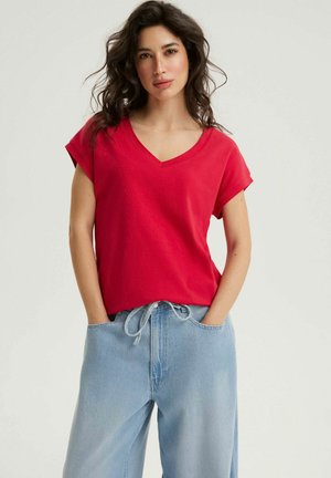 Kobieta o ciemnych, falowanych włosach, ubrana w czerwoną koszulkę z dekoltem w serek i jasnoniebieskie, szerokie dżinsy, z rękami w kieszeniach.