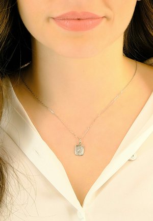 Collier en argent avec un pendentif rectangulaire, présentant un design détaillé, suspendu à une chaîne délicate, porté sur un chemisier blanc.