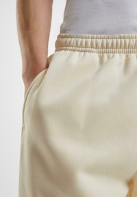 Primer plano de una persona que lleva pantalones de cintura elástica beige con las manos en los bolsillos laterales y una camiseta sin mangas blanca de canalé.