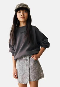Ragazza giovane che indossa un felpa grigio scuro, una gonna in denim acid wash con bottoni frontali e un cappellino a stampa leopardata, in piedi con una mano in tasca.