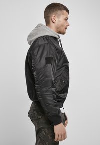 Jachetă bomber neagră cu fermoar în față, manșete elastice și un hanorak cu glugă gri dedesubt. Include buzunare laterale și o patch-uri cu velcro.