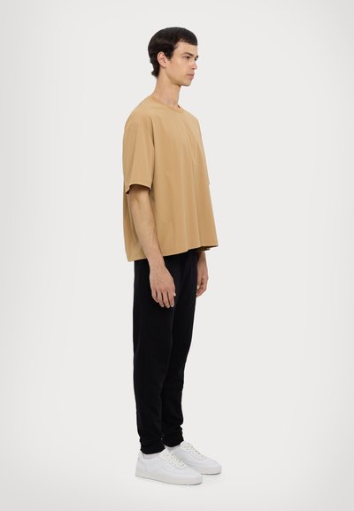 Chemise beige oversize à manches courtes avec un col rond, associée à un pantalon noir fuselé et des baskets blanches, présentée en profil latéral.
