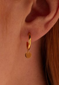 Boucle d'oreille en or avec un petit charme circulaire suspendu, présentant une surface lisse et brillante et un design minimaliste.