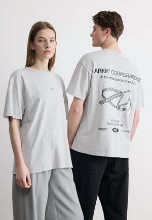 Zwei Personen tragen hellgraue T-Shirts der ARKK Corporation; eine zeigt die Vorderseite, die andere die Rückseite mit Text und Logo.