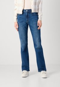 Kvinne iført blå vide jeans og hvite sneakers, kombinert med en lys stripete cardigan, stående foran en nøytral bakgrunn.