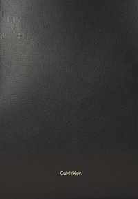 Surface en cuir noir texturé avec un petit logo Calvin Klein doré centré près du bas.