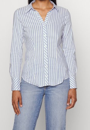 Femme portant une chemise blanche à manches longues avec des rayures verticales bleues et un jean bleu clair, debout devant un fond uni.