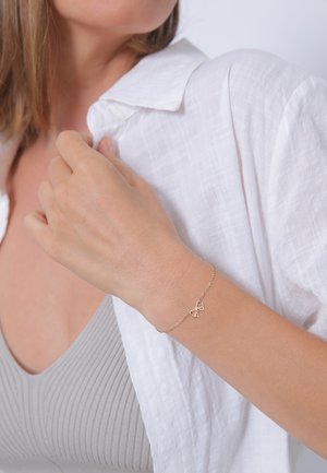 Zarter Armband aus Roségold mit einem schleifenförmigen Anhänger, der eine dünne Kette und eine elegante Textur aufweist, getragen an einem Handgelenk mit heller Haut.