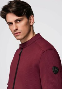 Herren Jacke in Burgunderrot mit strukturiertem Design, hohem Kragen, Frontreißverschluss und schwarzem Logo-Patch auf dem linken Ärmel.