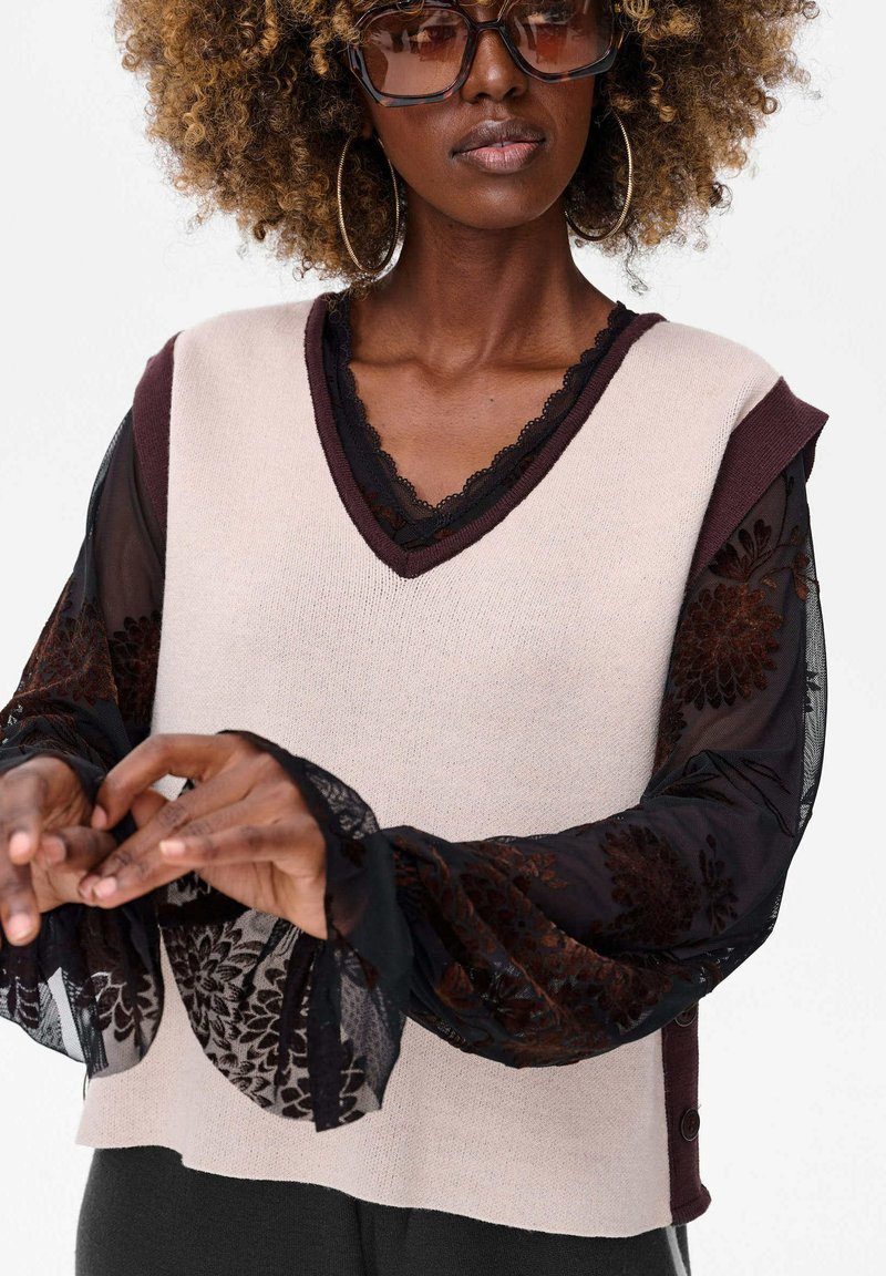 Beżowy dzianinowy top na ramiączkach z burgundowym wykończeniem, nałożony na przezroczystą czarną bluzkę w kwiatowy wzór z szerokimi koronkowymi mankietami. Całość zdobią akcenty guzikowe.