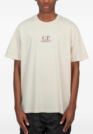 Uomo che indossa una maglietta beige oversize con la scritta "C.P. Company" sul petto e pantaloni neri a taglio largo su sfondo bianco.