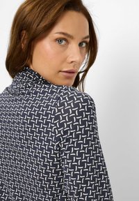 Una donna con gli occhi blu e i capelli castani si volta verso di spalle, indossando una maglietta blu navy con un motivo geometrico bianco su uno sfondo neutro.