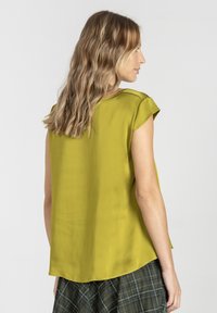 Kobieta z falującymi blond włosami, ubrana w limonkową, bezrękawnikową bluzkę i ciemnozieloną spódnicę w kratkę, stojąca bokiem na jednolitym tle.