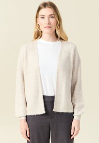 Ljus beige stickad cardigan med en mjuk textur, öppen front och långa ärmar, tillsammans med en vit topp och mörka byxor.