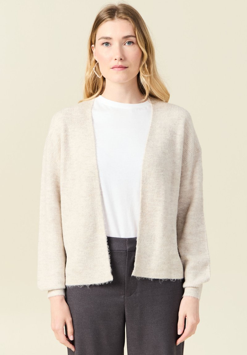 Ljus beige stickad cardigan med en mjuk textur, öppen front och långa ärmar, tillsammans med en vit topp och mörka byxor.