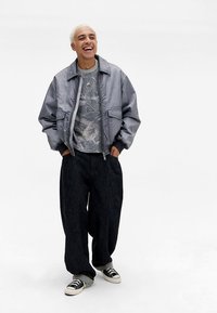 Pilkas bomberio stiliaus švarkas su lygiu paviršiumi, derinamas su raštuotu pilku džemperiu ir laisvomis juodomis džinsinėmis kelnėmis. Juodi sportbačiai užbaigia įvaizdį.