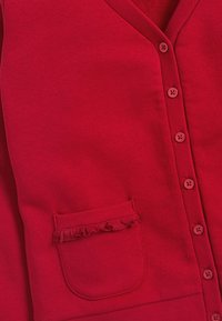 Un cardigan rouge avec un col en V, une fermeture à boutons, un tissu texturé et une poche ornée de volants. Texture lisse et douce avec des accents côtelés.