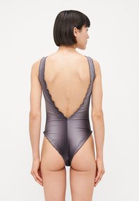 Pilkos spalvos vientisas maudymosi kostiumėlis su banguota iškirpte ir žema nugara, pagamintas iš lygaus, elastingo audinio. Kirpimas suteikia prigludusią siluetą.