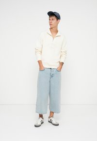 Felpa bianca con zip e polsini a costine, jeans a gamba larga di un blu chiaro e sneakers nere/grigie, completati da un berretto blu scuro.