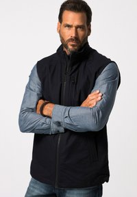 Schwarz ärmellose Weste mit Reißverschluss, getragen über einem hellblauen Langarmhemd. Verfügt über eine glatte Textur und eine lockere Passform.