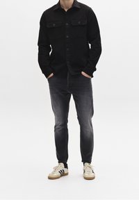 Man som bär en svart knapp-up skjorta, blekta svarta jeans och vita sneakers med svarta ränder, står med händerna i fickorna.
