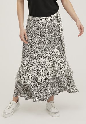 Kobieta ubrana w czarny top, białe sneakersy i warstwową czarno-białą midi spódnicę w kwiatowe wzory i kropki.