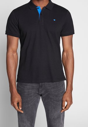 Mężczyzna ubrany w czarną koszulkę polo z krótkim rękawem z niebieskim detalem przy kołnierzyku i małym niebieskim logo, w połączeniu z wyblakłymi czarnymi dżinsami.