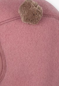 Kogumüts pehmes roosas villases materjalis, mille peal on ümmargune pruun pom-pom aktsent. Sile tekstuur ja nähtav tikkimine serva ääres.