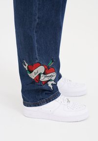 Dark blue denim jeans featuring a prominent red heart embroidered design with black outlines and a white ribbon inscribed with "Ed Hardy." White sneakers.