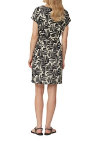 Kurzärmliges Kleid in Creme mit schwarzem Blätterdruck, tailliert. Es hat eine gerade Form und endet oberhalb der Knie, kombiniert mit schwarzen Sandalen.