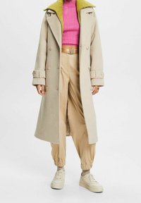 Henkilö, joka pitää beigeä pitkää takkia, jossa on nappiyksityiskohtia, vaaleanpunaista ribbikuvioista lyhytvartista toppia, beigejä vyöllisiä housuja, jotka ovat kireät nilkoista, beigejä lenkkareita ja harmaita sukkia.