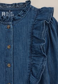 Denimová blůza v tmavě modré barvě s volánkovým límcem a rukávy, zdobená dřevěnými knoflíky. Tkanina má jemnou textura a uvolněný design.