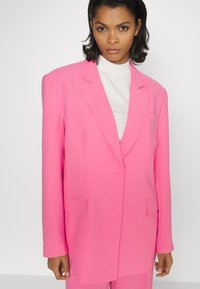 Blazer rosa feito de tecido suave, apresentando um design ajustado, lapelas recortadas e bolsos frontais. As mangas longas chegam até os pulsos.