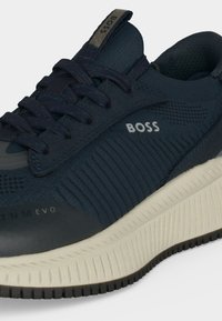 Σκούρο μπλε αθλητικό παπούτσι BOSS με υφασμάτινο επάνω μέρος με υφή, μαύρα κορδόνια και παχύ λευκό πέλμα με ορατό μαύρο πέλμα.