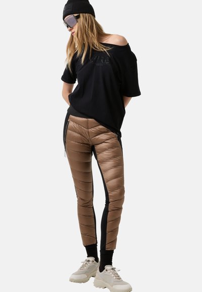 Brune polstrede bukser med sorte sidepaneler, parret med en løs sort t-shirt og kraftige hvide sneakers. Teksturen er glat og skinnende.