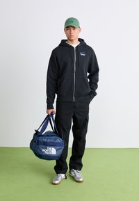 Moški v črni kapuci, črnih cargo hlačah, z zeleno kapo, drži modro torbo The North Face in stoji na zeleni tleh ob beli steni.