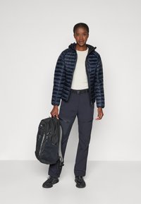 Marineblauwe pufferjas met een capuchon, lichte shirt, donkere cargobroek en een zwarte rugzak, met ritsen en textuur details.