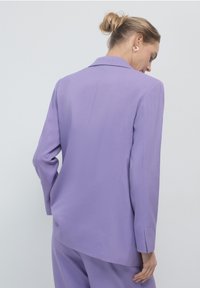 Blazer lilla con un design su misura, revers a punte e una finitura testurizzata. Le maniche sono lunghe con un leggero dettaglio di risvolto.