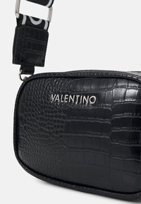 Borsa a tracolla in pelle nera con texture a coccodrillo, logo "Valentino" in argento e tracolla regolabile con motivo a contrasto. Forma rettangolare.