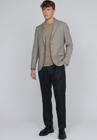 Randig blazer i beige och svart tyg, kombinerad med en ljusbrun skjorta, svarta plisserade byxor och matchande svarta skor.