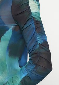 Plavi i zeleni tie-dye efekt, skupljena dugo rukava, prilagođeni materijal s vidljivim šavovima i nabranom teksturom na rukavu.