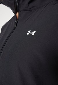 Črna zip-up jakna izdelana iz mehkega, raztegljivega blaga. Ima povišan ovratnik in velik bel logotip na prsih. Vidna zadrga za prilagajanje.