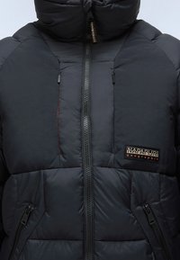 Svart puffjacka med hög krage, dragkedja framtill och fickor, quiltat tyg, med en logotyp på vänster sida.