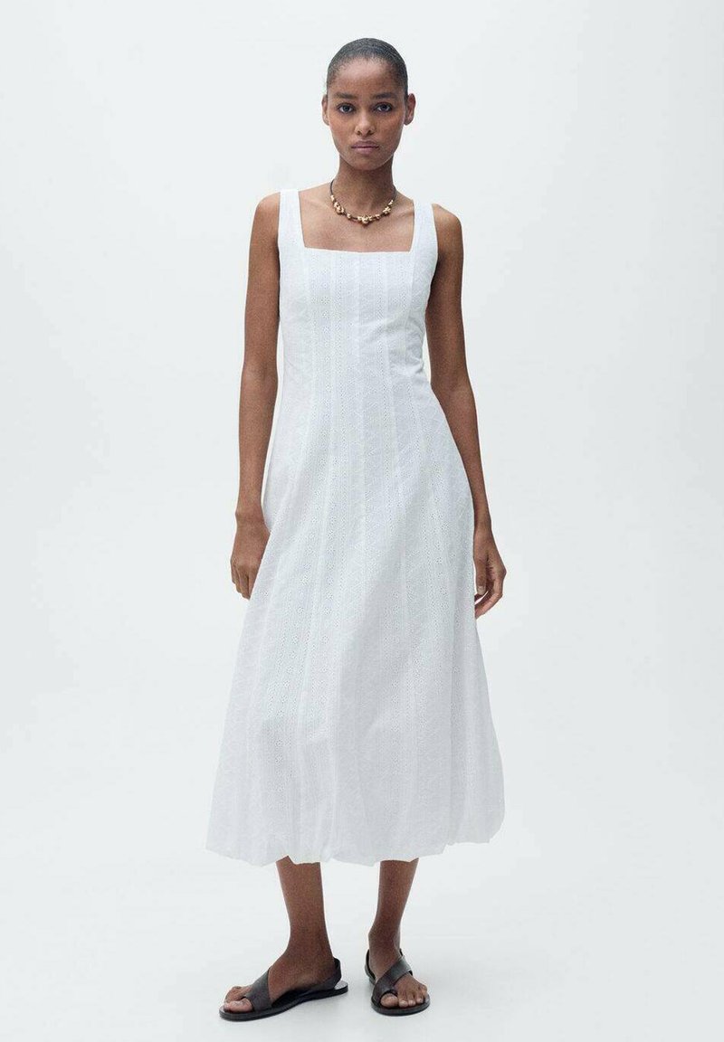 Vit ärmlös klänning med fyrkantig halsringning, knälång, med broderade detaljer och ett flödande, texturerat tyg. Bärs med svarta sandaler.