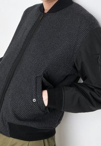 Veste bomber avec un corps en mélange de laine gris texturé, des manches en tissu noir, des poignets côtelés et des poches zippées.