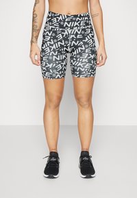 Henkilö, joka on pukeutunut musta-valkoisiin Nike-logoilla varustettuihin polkupyöräshortseihin ja mustiin Nike Air -lenkkareihin, seisoo vaaleaa taustaa vasten.