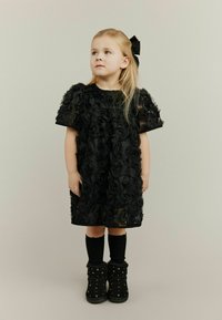 Crna haljina s 3D aplikacijama u obliku ruža, kratkim rukavima i okruglim izrezom. Uparena s crnim čarapiam do koljena i ukrašenim gležnjačama.