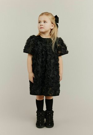 Svart klänning med 3D-rosapplikationer, korta ärmar och rund halsringning. Kombinerad med knähöga svarta strumpor och dekorerade ankellånga stövlar.
