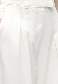 Κοντινό πλάνο λευκού παντελονιού με ψηλή μέση, μπροστινές πιέτες, πλαϊνές τσέπες και ορατούς θηλιές ζώνης στη μέση ενός ατόμου.